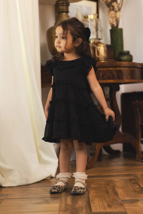 A young girl with dark hair stands in a room wearing a black ruffled tiered dress, white ruffled socks, and the Noralee Alice Bow | Black