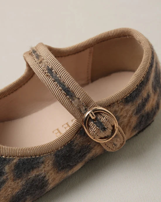 Close-up of a Mary Jane flat shoe featuring a brown and black leopard print pattern, a beige ribbed elastic strap, and a gold-tone metal ring buckle.