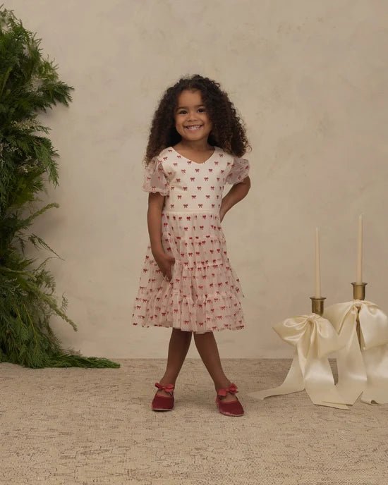 A young girl with curly dark hair stands in a studio setting wearing a short-sleeve pink dress with a red pattern, paired with red shoes. The background features a textured beige wall, a green plant on the left, and decorative cream ribbons and candle holders on the right.