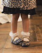 A close-up of a child's legs wearing leopard-print baby shoes with white ruffled socks, standing on a wooden floor.