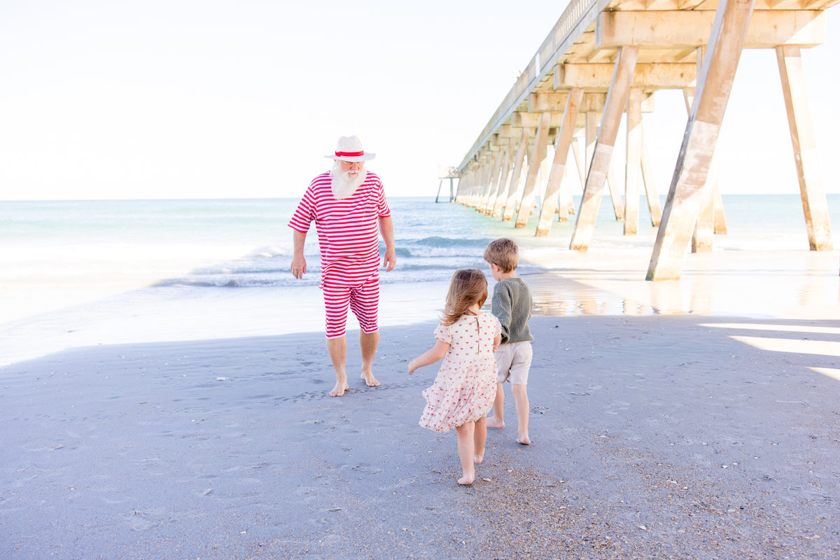 A man in a red and white striped shirt and shorts walks on a sandy beach with two children under a wooden pier.