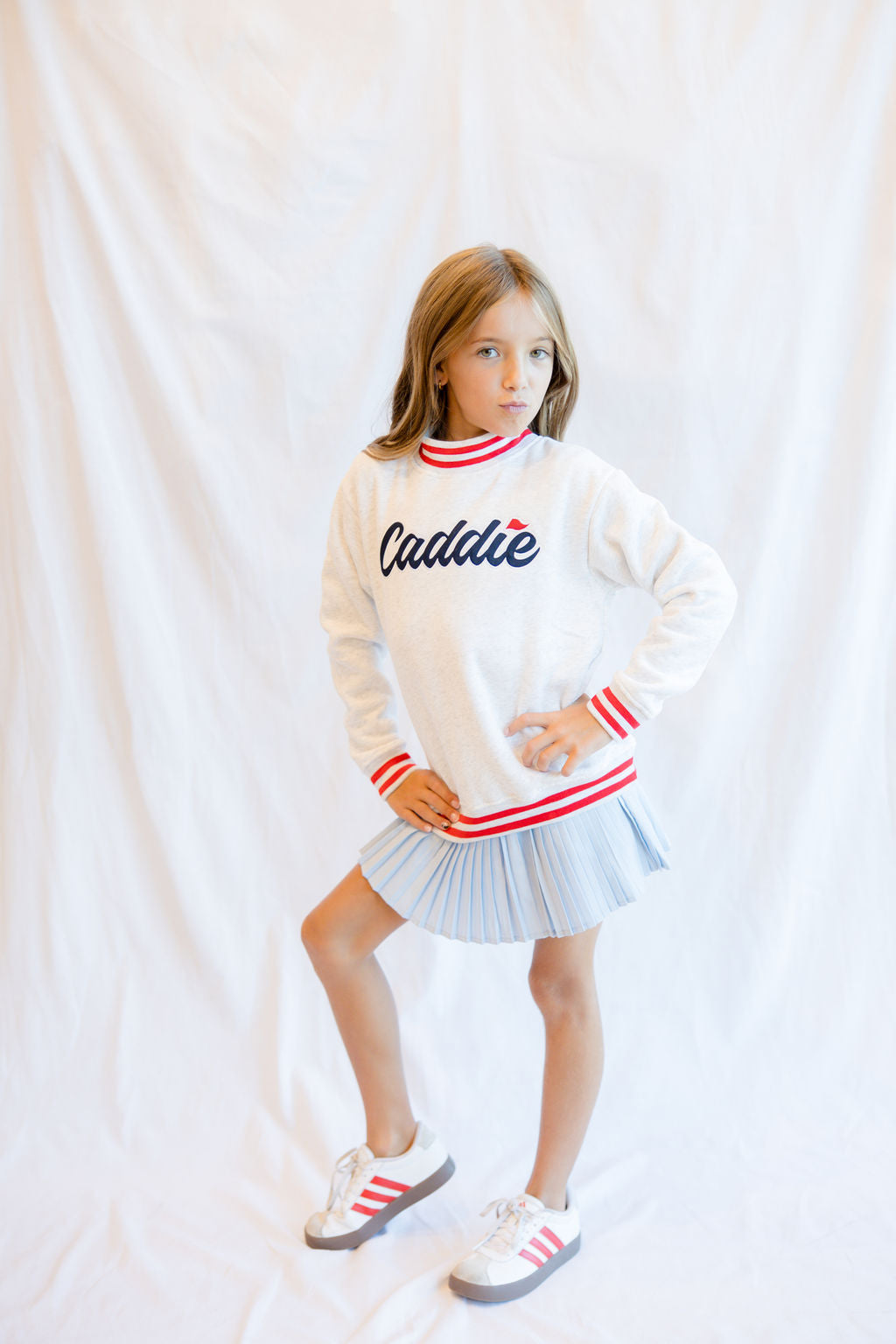 A young girl modeling a white long-sleeve sweatshirt with 'Caddie' written on the chest in navy script with a red dot, paired with a light blue pleated mini skirt and white sneakers with red stripes, posed against a white fabric backdrop.