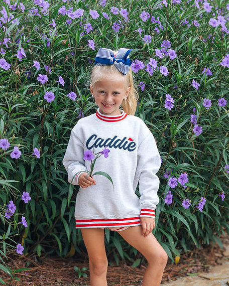 A young girl stands outdoors in front of a dense background of green plants and purple flowers, wearing a white crewneck sweatshirt with red and navy blue trim and the name 'Caddie' printed on the front.