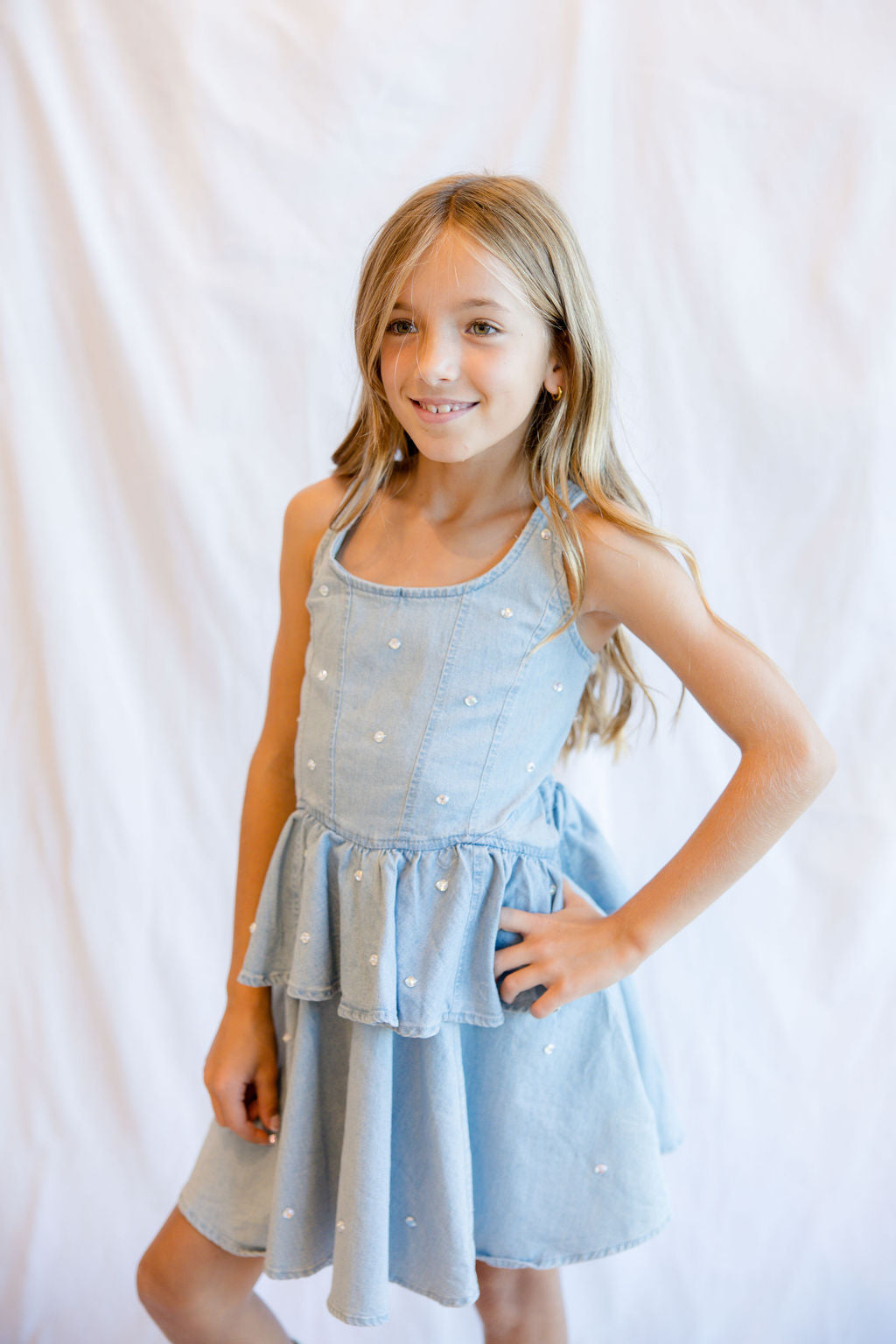 A young girl with blonde hair wearing a light blue denim-style sleeveless dress with white embroidered dots, posing against a white fabric backdrop.