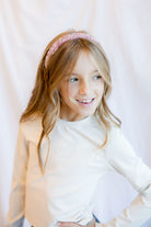 A young girl with long light brown hair wearing a pink glittery headband and a white long-sleeve top, posing against a soft white fabric backdrop.