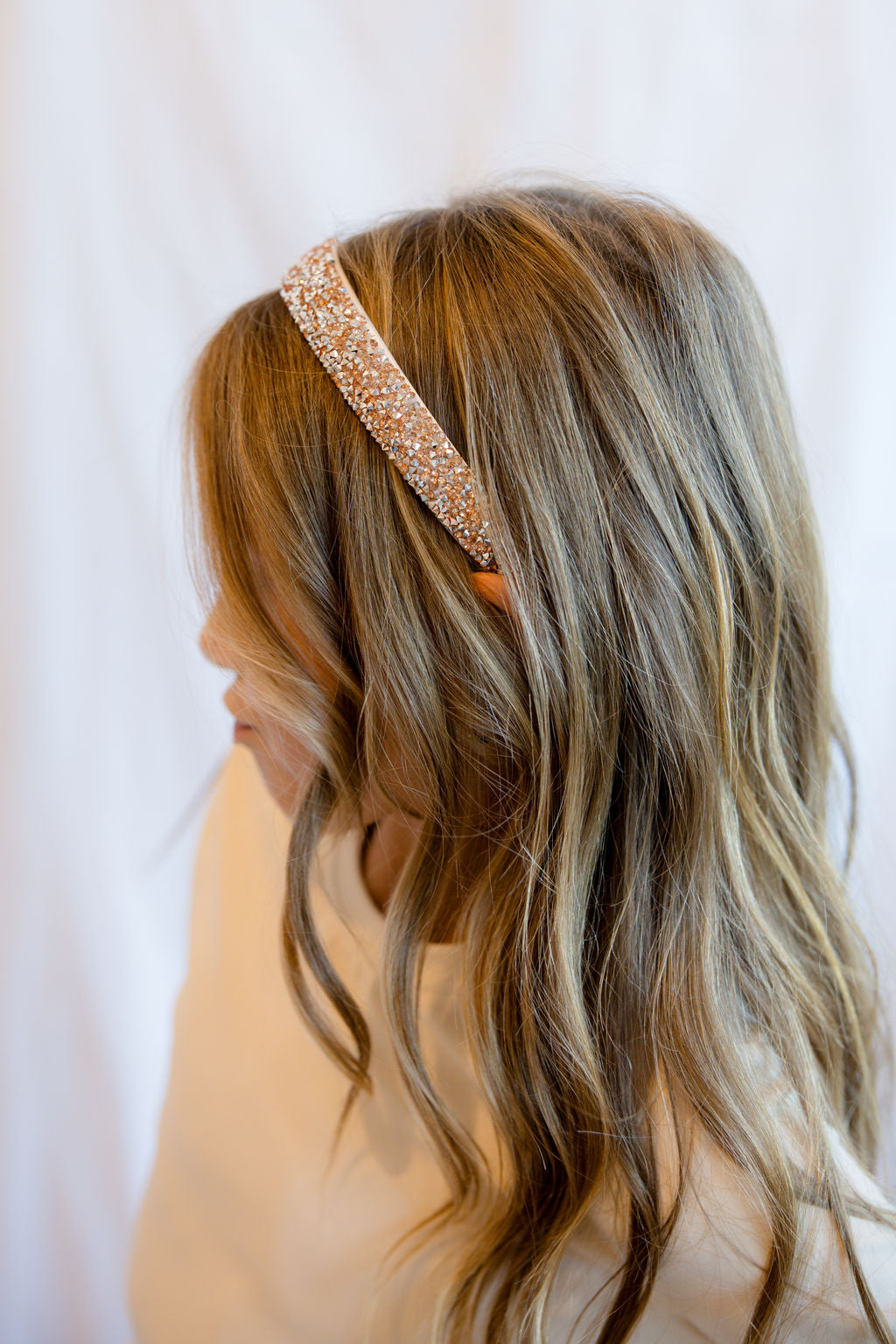 A woman shown from the side and back wearing a sparkling rose gold headband with a small orange bow accent, set against a soft white draped background.