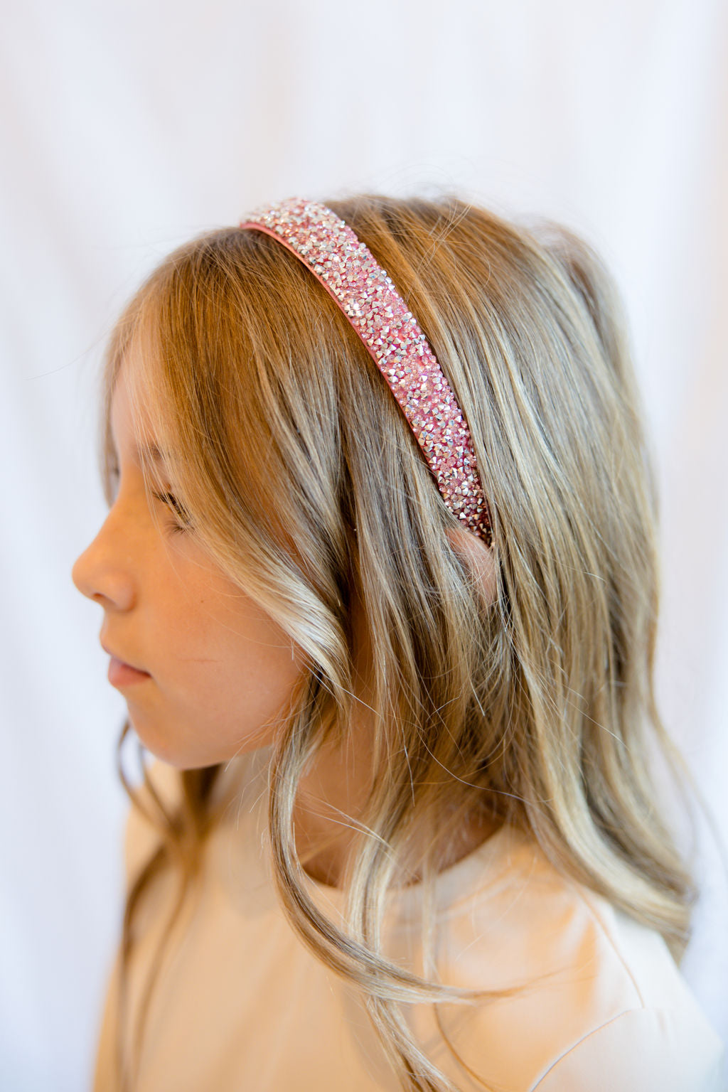 A young girl with blonde hair wearing a sparkling pink glitter headband, shown in profile against a soft white background.