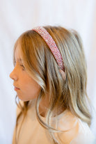 A young girl with blonde hair wearing a sparkling pink glitter headband, shown in profile against a soft white background.