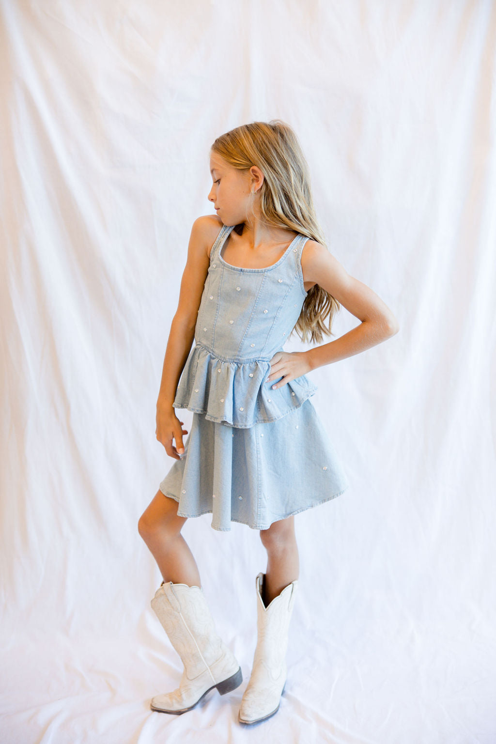 A young girl with blonde hair stands in profile wearing a light blue sleeveless dress with a ruffled peplum hem and white cowboy boots, posed against a white fabric backdrop.