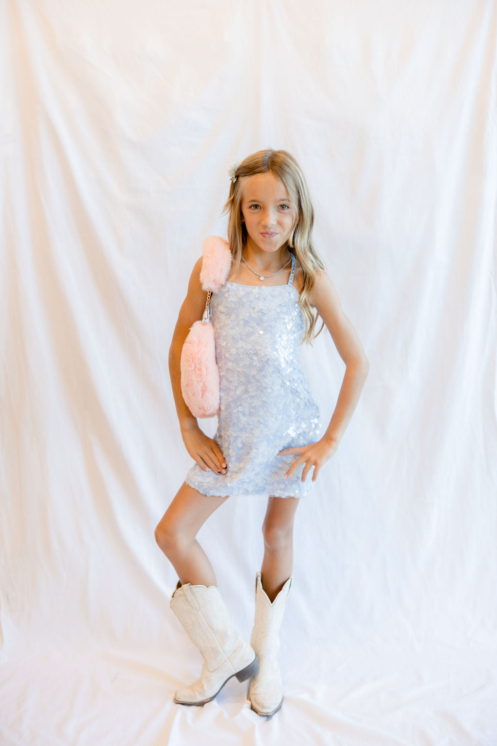 A young girl stands against a white draped fabric backdrop, wearing a sparkly silver sequin mini dress, white cowboy boots, and holding a pink fuzzy accessory.