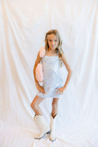 A young girl stands against a white draped fabric backdrop, wearing a sparkly silver sequin mini dress, white cowboy boots, and holding a pink fuzzy accessory.