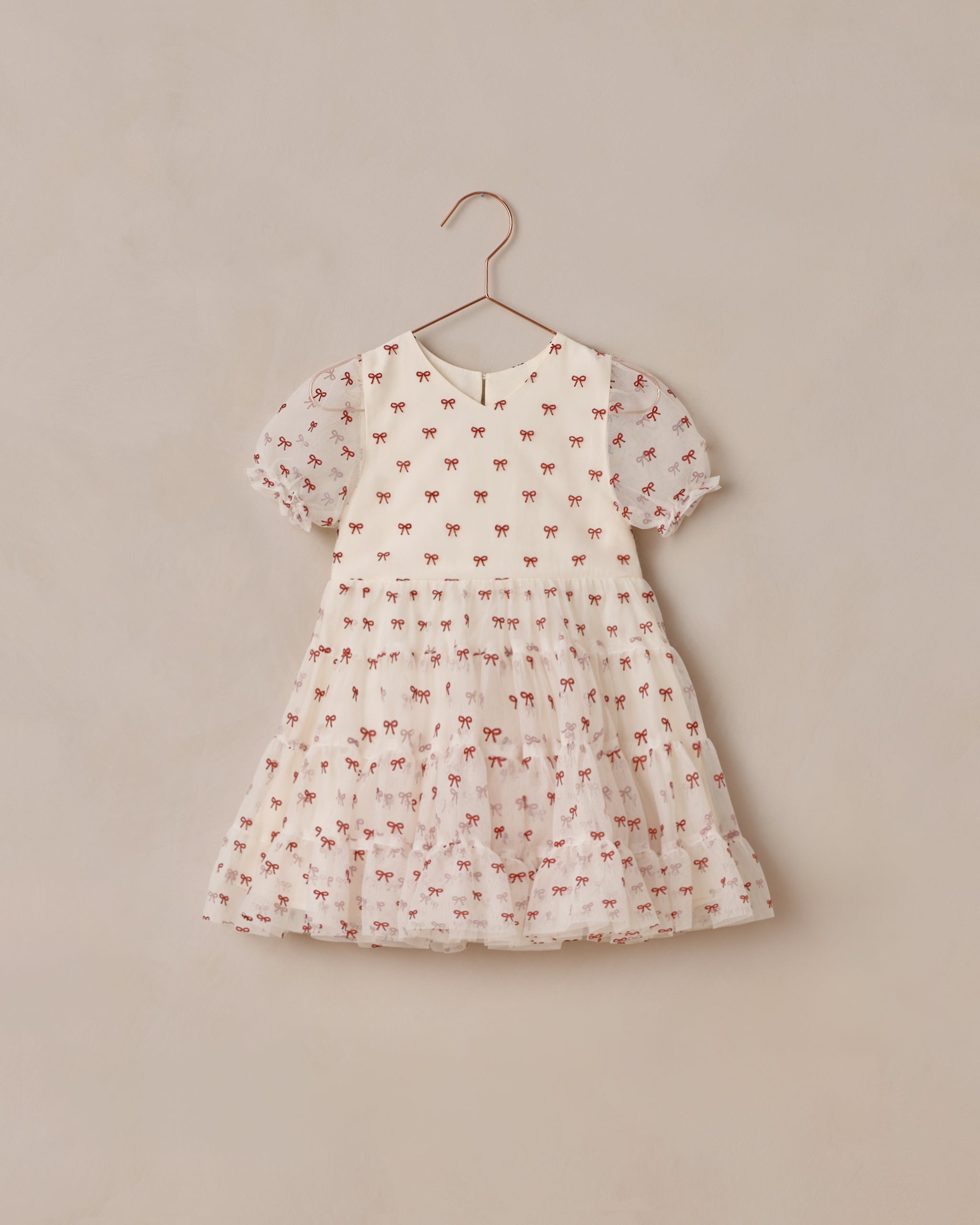 A short-sleeve baby dress in a warm white fabric, decorated with a delicate all-over print of small red bows. The dress is shown hanging on a simple metal hanger. Noralee Dottie Dress | Ruby Bows