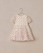 A short-sleeve baby dress in a warm white fabric, decorated with a delicate all-over print of small red bows. The dress is shown hanging on a simple metal hanger. Noralee Dottie Dress | Ruby Bows