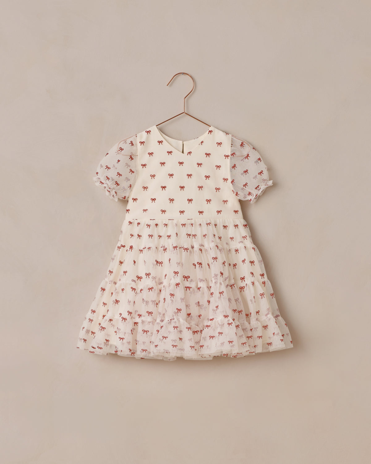 A short-sleeve baby dress in a warm white fabric, decorated with a delicate all-over print of small red bows. The dress is shown hanging on a simple metal hanger. Noralee Dottie Dress | Ruby Bows