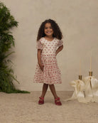 A young girl with curly dark hair stands in a studio setting wearing a short-sleeve pink dress with a red pattern, paired with red shoes. The background features a textured beige wall, a green plant on the left, and decorative cream ribbons and candle holders on the right.