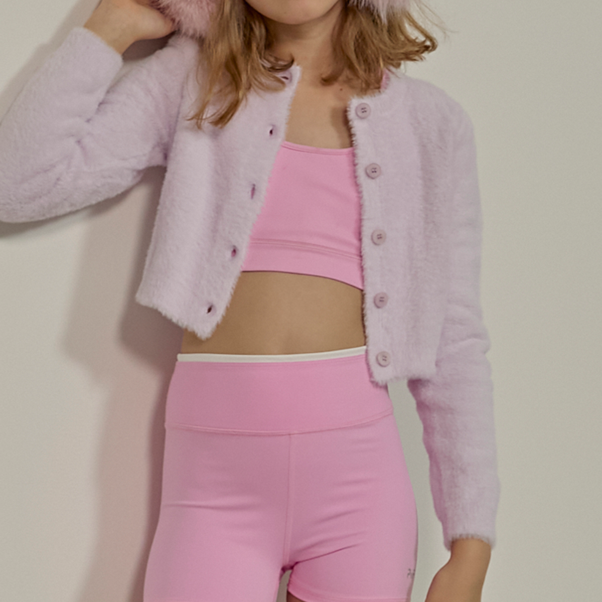 A model wearing a light pink fuzzy cropped cardigan with matching buttons, a pink sports bra, and pink leggings against a plain beige wall.