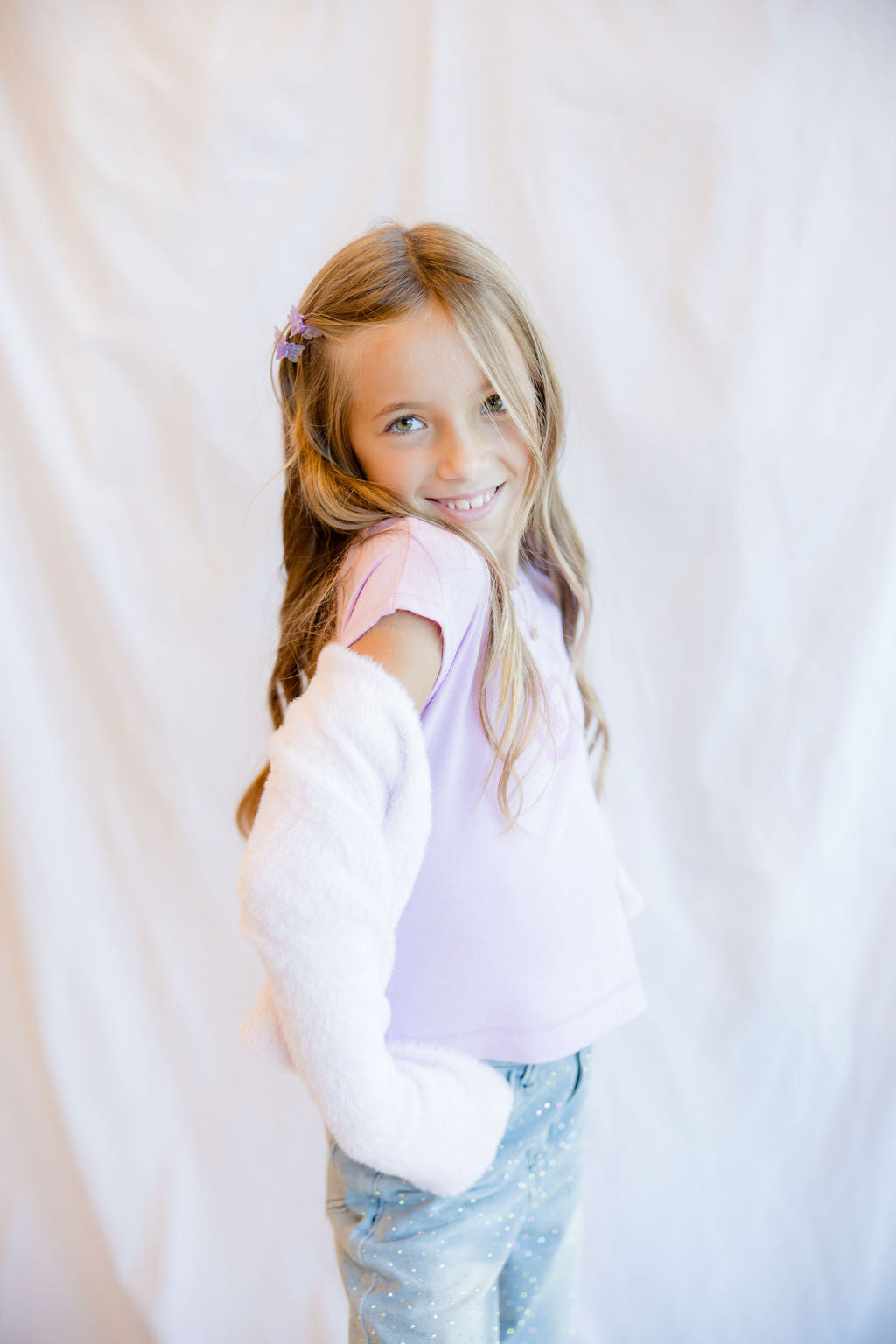 A young girl with long blonde hair looks back over her shoulder while wearing a light pink top, a white cardigan, and light blue jeans, standing against a white fabric backdrop.