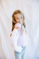 A young girl with long blonde hair looks back over her shoulder while wearing a light pink top, a white cardigan, and light blue jeans, standing against a white fabric backdrop.
