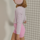 A woman shown from the side wearing a fuzzy light pink cropped cardigan, a matching fuzzy pink beanie, and pink fitted shorts against a white tiled wall.