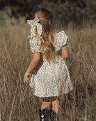A young girl with long blonde hair is seen from behind, wearing a cream-colored dress with a small floral pattern and a large matching hair bow, standing in a field of dry grass.
