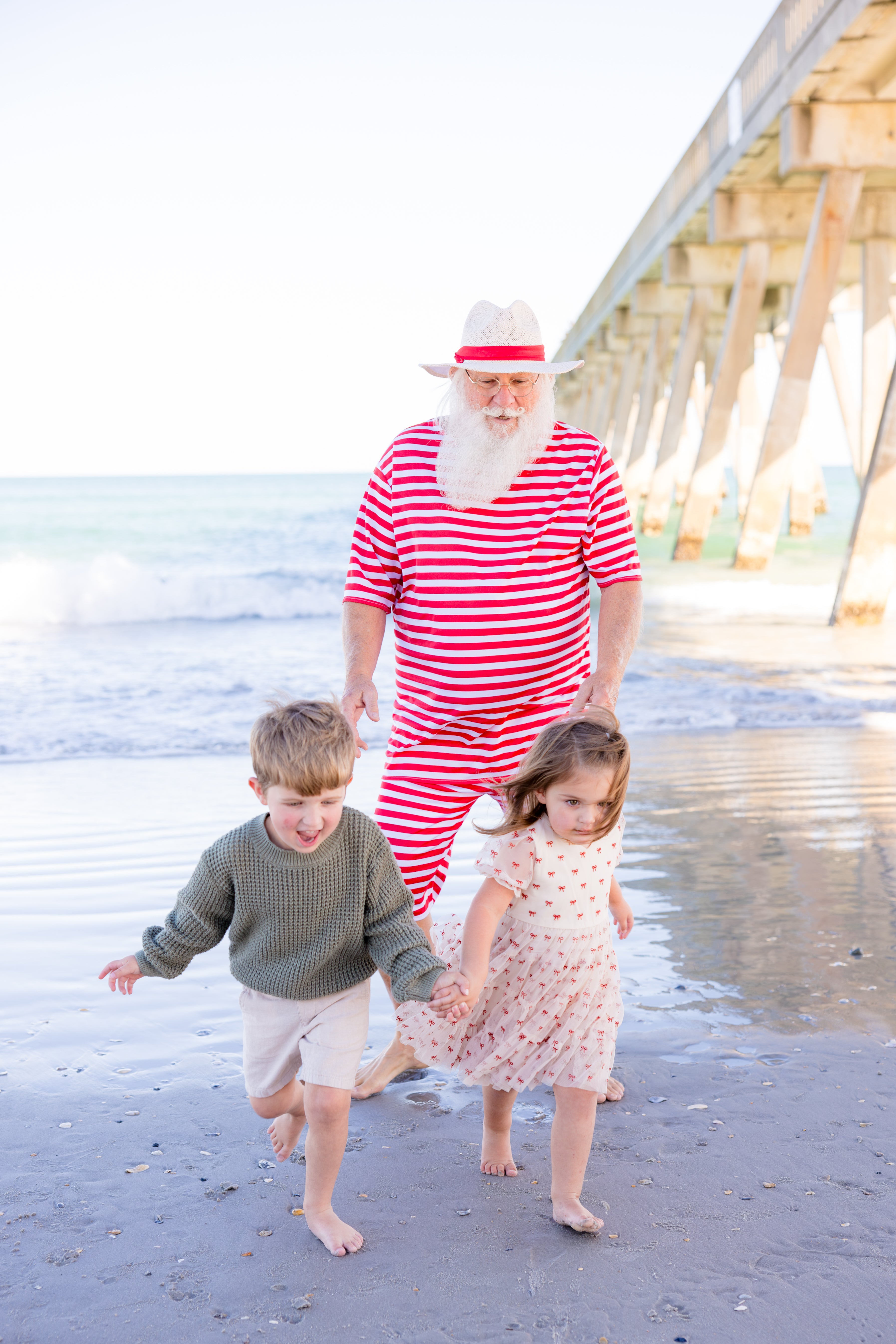 Santa Claus wearing a red and white striped outfit and a white hat with a red band, walks barefoot on a beach with two young children holding hands. A wooden pier is visible in the background.