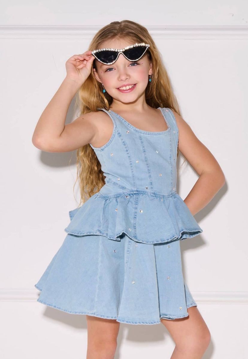 A young girl with long blonde hair models a light blue denim tiered dress with rhinestone embellishments. She is wearing white-framed sunglasses with rhinestones and is posing in front of a white wall with molding.